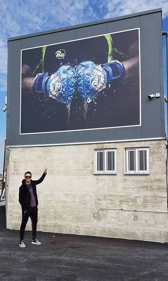 Ramiro Gonzales en tenue décontractée, portant des lunettes de soleil, pointe du doigt une immense affiche publicitaire sur un mur extérieur. L’affiche représente une paire de gants de gardien de but RG bleus